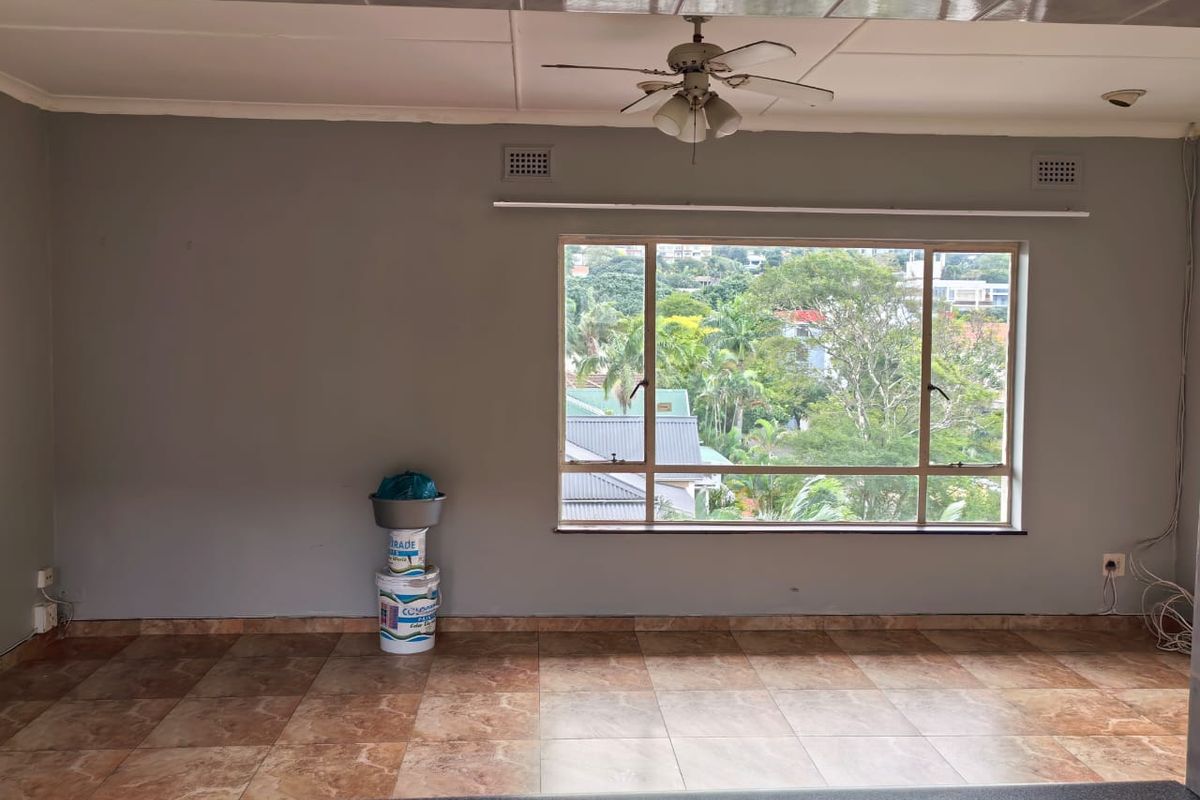 An empty living room featuring tiled flooring, a ceiling fan, and a large window with views of the surrounding neighborhood.