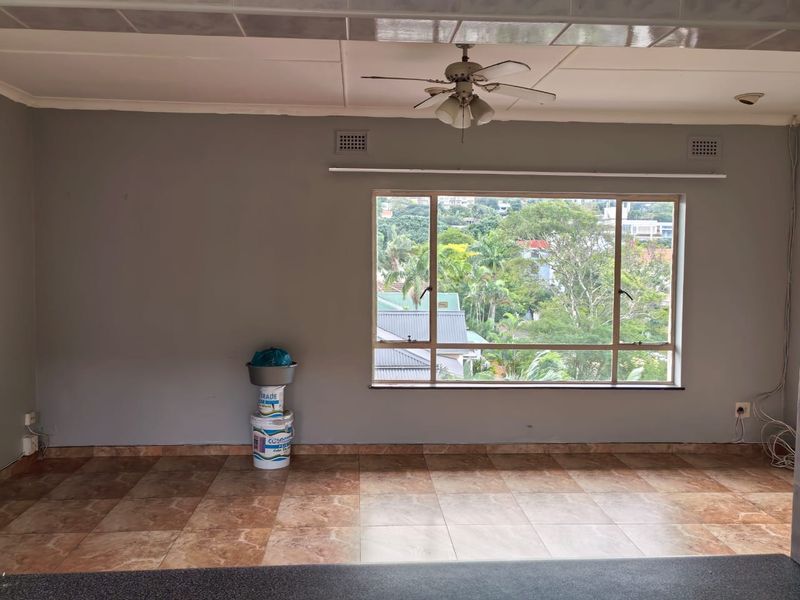 An empty living room featuring tiled flooring, a ceiling fan, and a large window with views of the surrounding neighborhood.