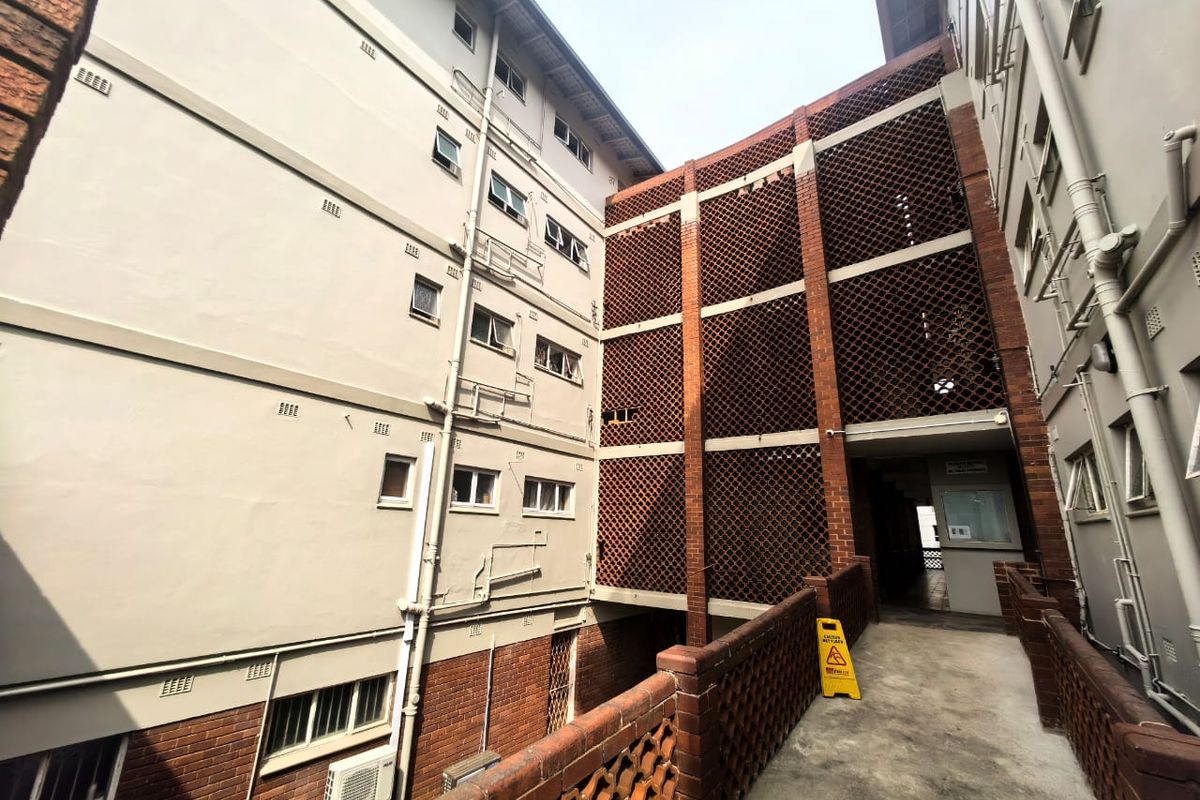 Exterior view of an apartment building complex featuring a concrete walkway, decorative brick screening, and exposed exterior infrastructure.