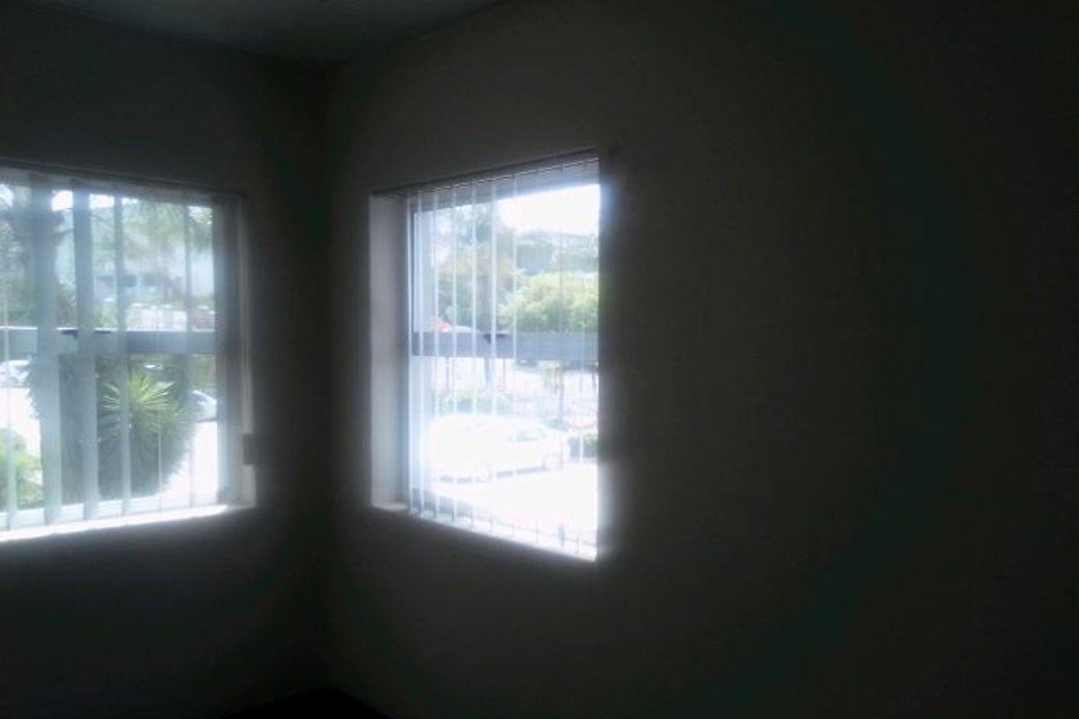 A vacant office space featuring large windows with vertical blinds and security bars, offering natural light and a view of the exterior.