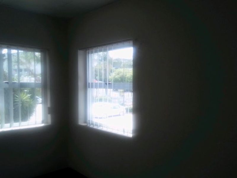 A vacant office space featuring large windows with vertical blinds and security bars, offering natural light and a view of the exterior.