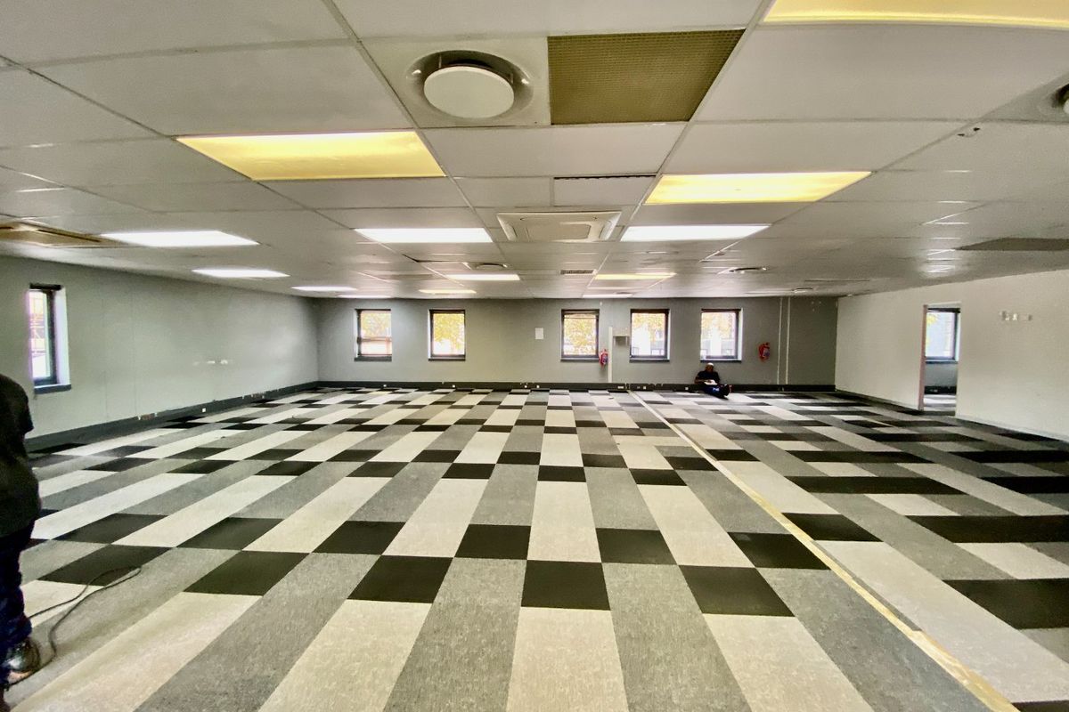 A large, open-plan commercial office space featuring a suspended ceiling, recessed lighting, and a patterned carpet tile floor.