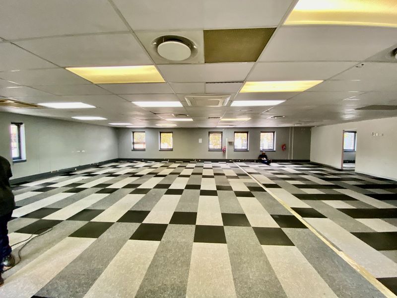A large, open-plan commercial office space featuring a suspended ceiling, recessed lighting, and a patterned carpet tile floor.