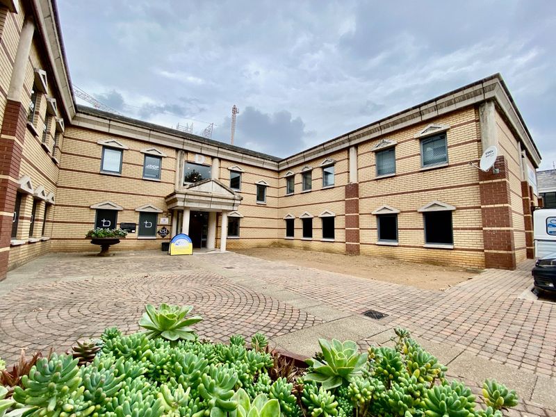 A commercial office building exterior featuring a brick facade, paved courtyard entrance, and multi-pane windows under a cloudy sky.