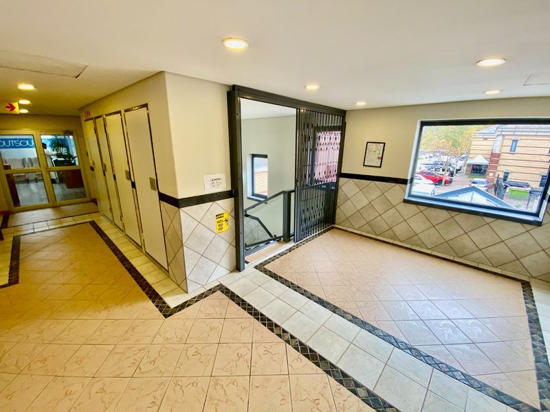 A bright, tiled commercial hallway featuring recessed lighting, a security gate leading to a staircase, and large windows providing natural light.