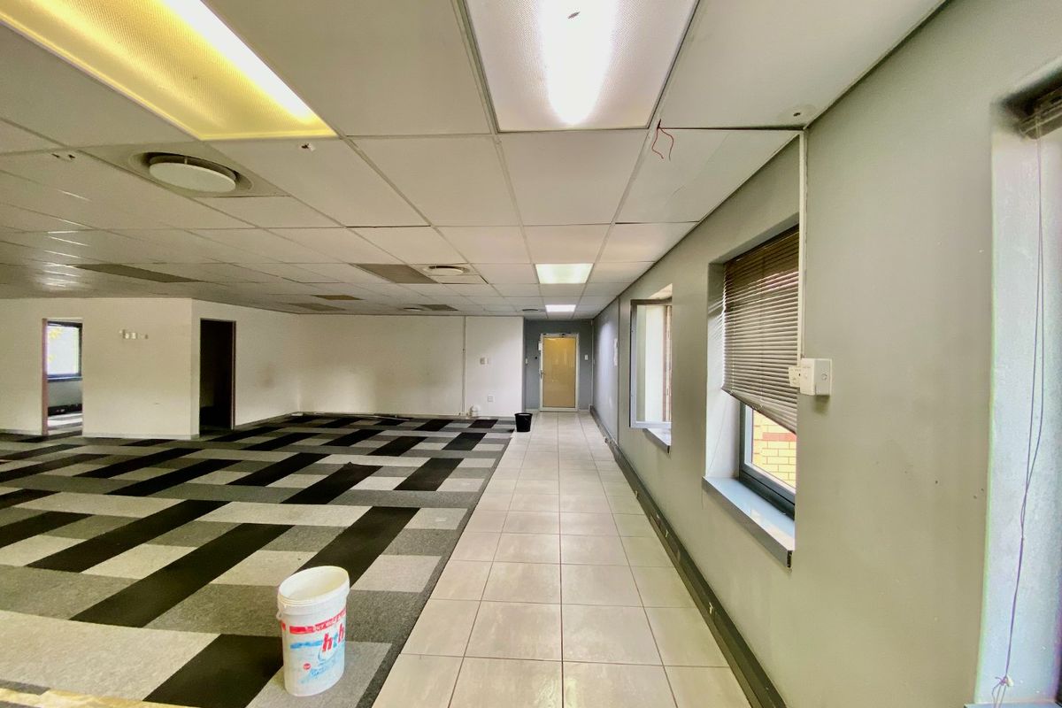 An open-plan commercial office space featuring a mix of carpet and tile flooring, suspended ceilings with recessed lighting, and large windows with blinds.