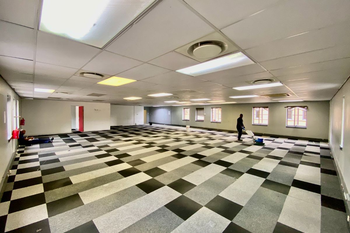 A spacious, open-plan commercial office area featuring a suspended ceiling, recessed lighting, air conditioning vents, and a checkered carpet tile floor.