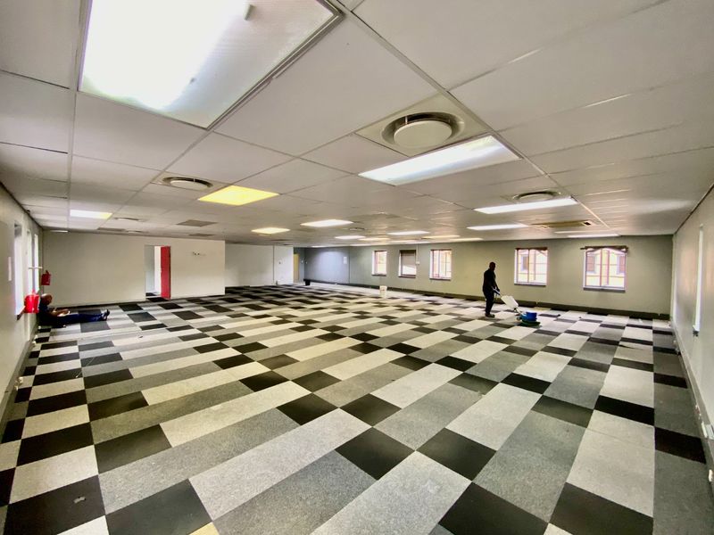 A spacious, open-plan commercial office area featuring a suspended ceiling, recessed lighting, air conditioning vents, and a checkered carpet tile floor.