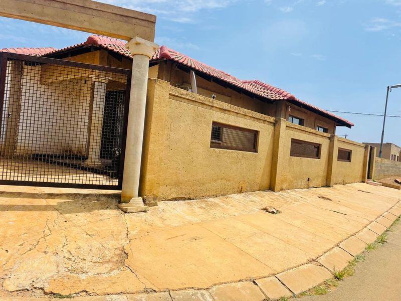 Front exterior view of the property featuring a perimeter wall, security gate, and tiled roof, showing signs of weathering and pavement cracking.