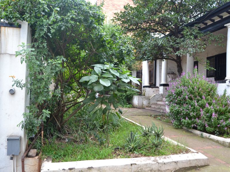 Front entrance area featuring a covered porch with classical columns, a paved walkway, and a raised garden bed requiring maintenance.