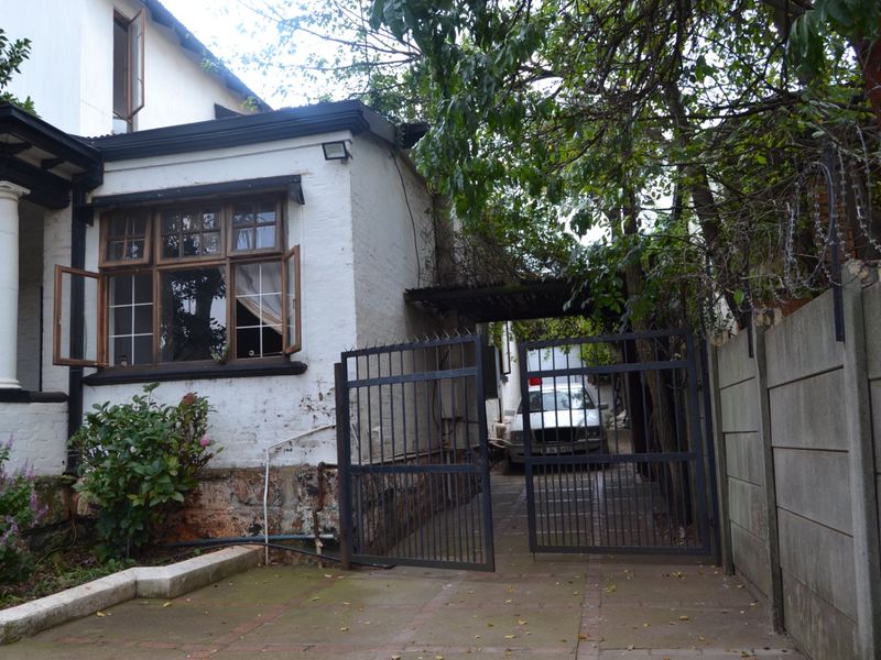 Front exterior view showing a paved driveway, metal security gate, precast boundary wall, and the side facade of the house with timber-framed windows.