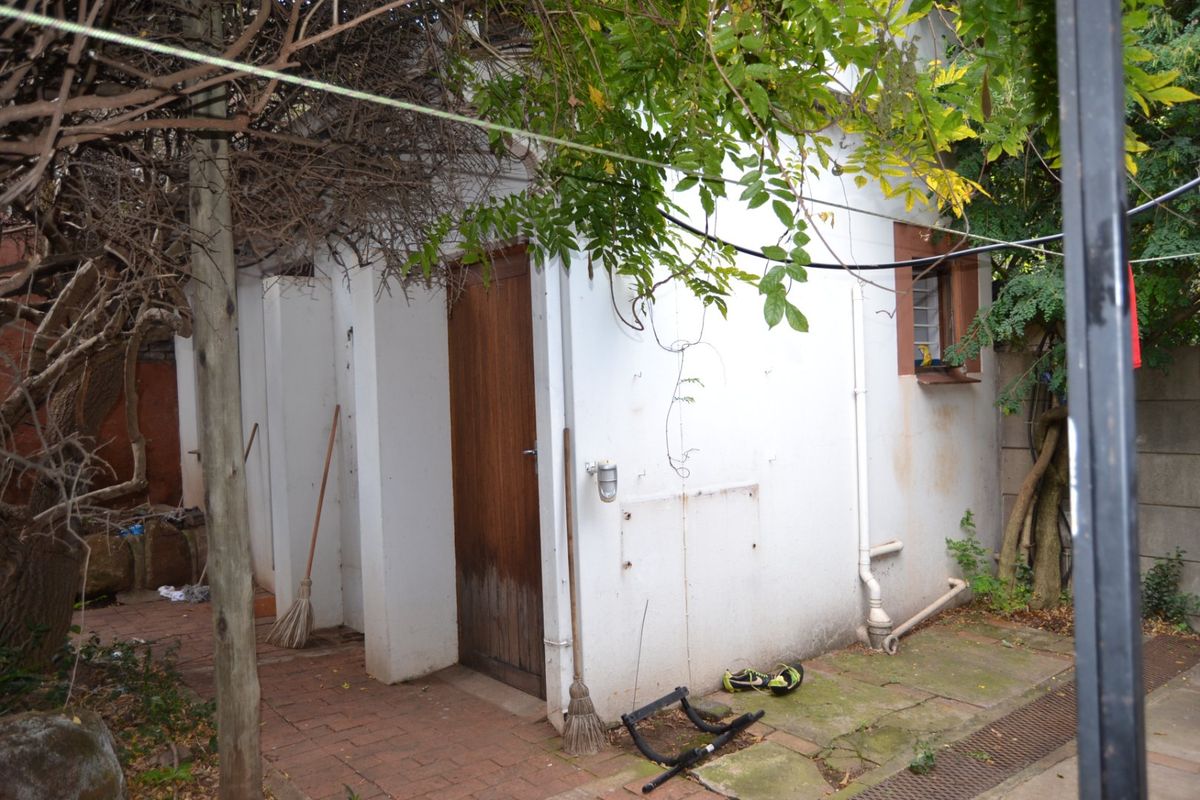 Rear exterior view of a utility structure featuring brick paving, a timber door, and external plumbing fixtures in need of maintenance.