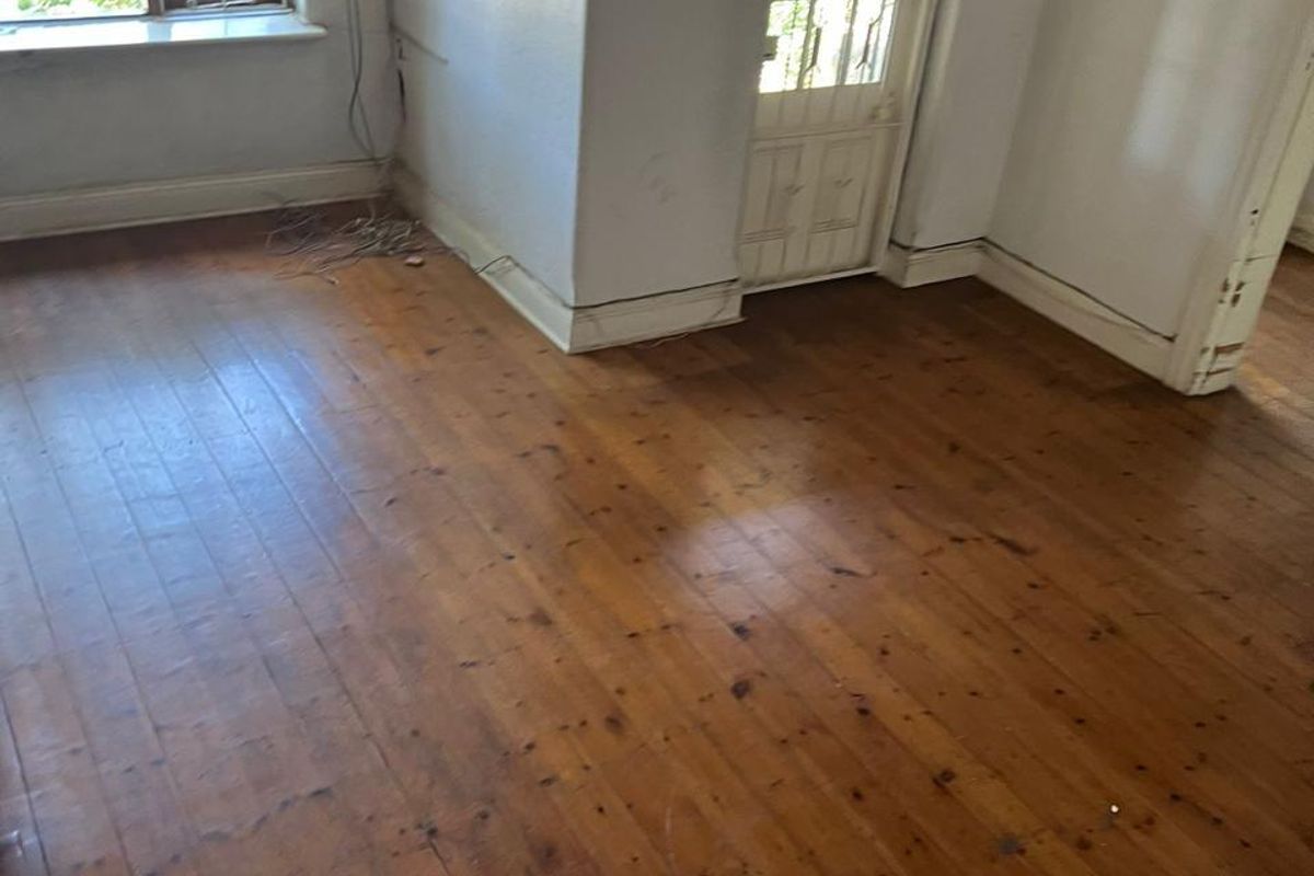 An empty living room featuring original timber flooring, mullioned windows, and a classic ceiling light fixture, requiring minor cosmetic updates to skirting and walls.