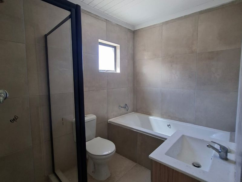 A functional bathroom featuring a walk-in shower with black frame, built-in bathtub, vanity unit, and neutral floor-to-ceiling tiling.