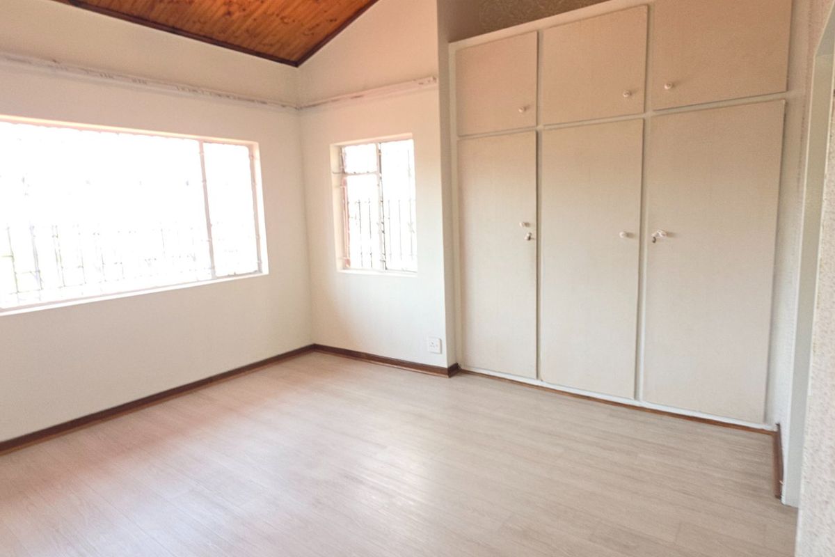 A bright, empty bedroom featuring built-in cupboards, laminate flooring, and a vaulted timber ceiling with security bars on the windows.