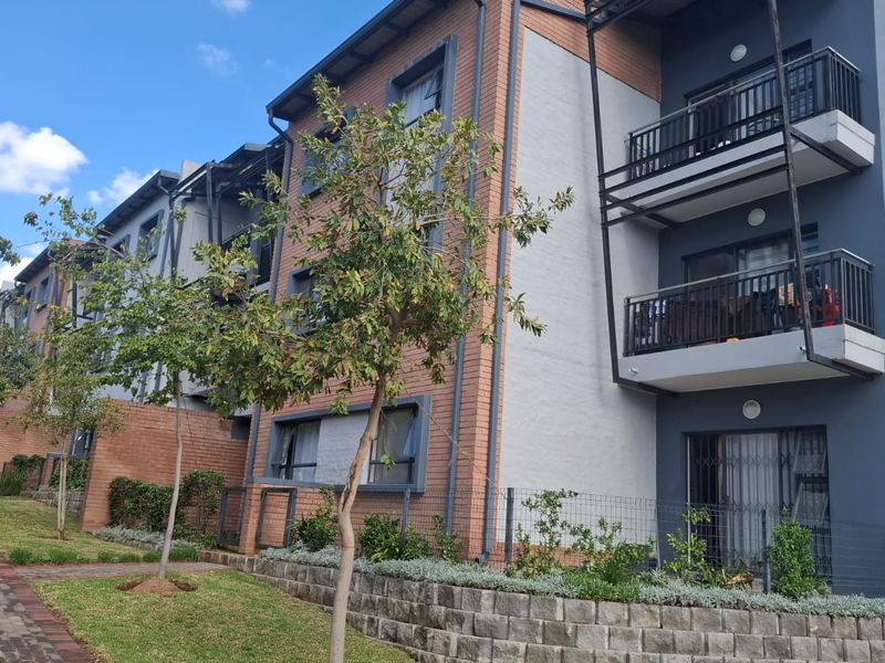 A view of the apartment building exterior featuring brick and rendered facades, private balconies with metal railings, and a landscaped garden area with paved walkways.