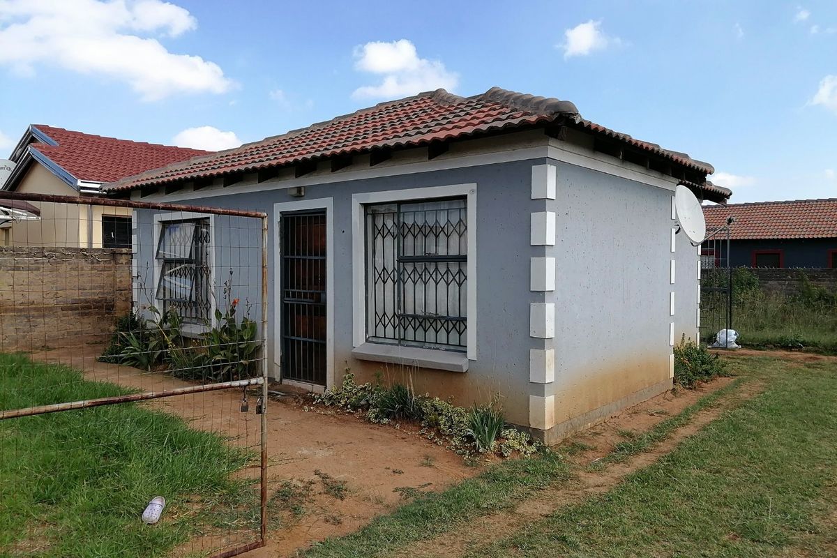 Rear view of a detached house featuring a tiled roof, security bars on windows, and a security gate, set within an unpaved garden area.