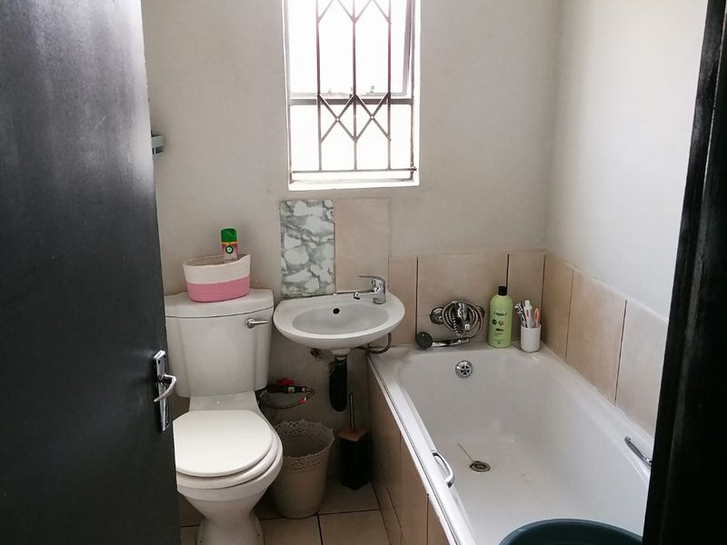 A compact bathroom featuring a standard bathtub, pedestal basin, and toilet with tiled finishes and a window with security bars.