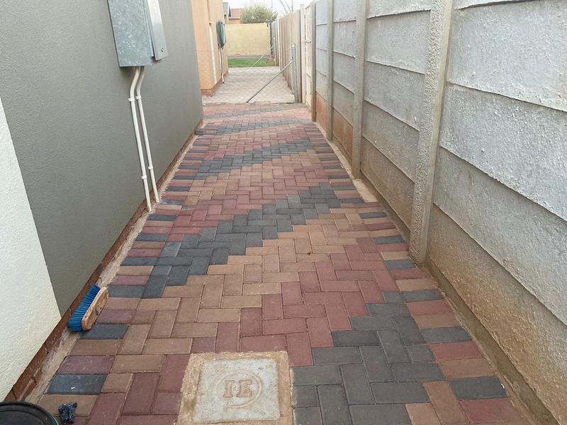 A clean, paved side walkway featuring a patterned brick design, bordered by a precast concrete boundary wall and the side of the house.