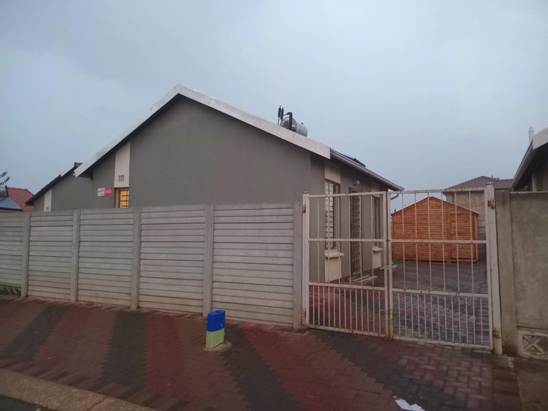 Front view of the property featuring a precast concrete boundary wall, metal security gate, paved driveway, and a detached timber garden shed.