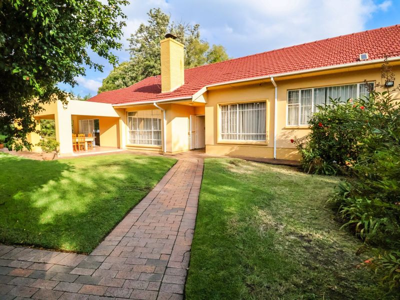 A well-maintained rear exterior view of the house featuring a red tiled roof, yellow facade, paved walkway, and a covered patio area.