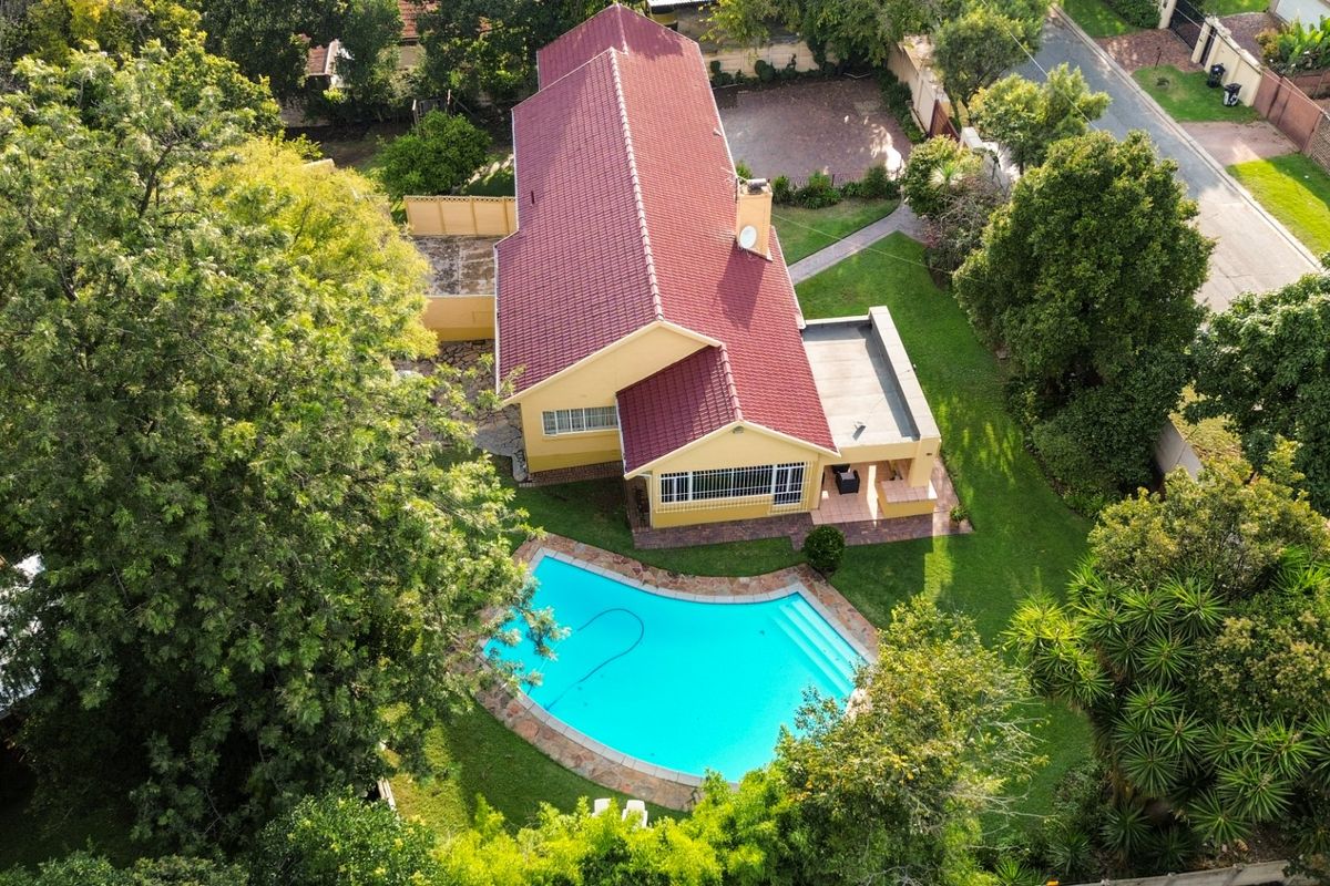 Aerial view of a residential property featuring a large swimming pool, expansive garden, paved patio area, and a well-maintained house with a tiled roof.