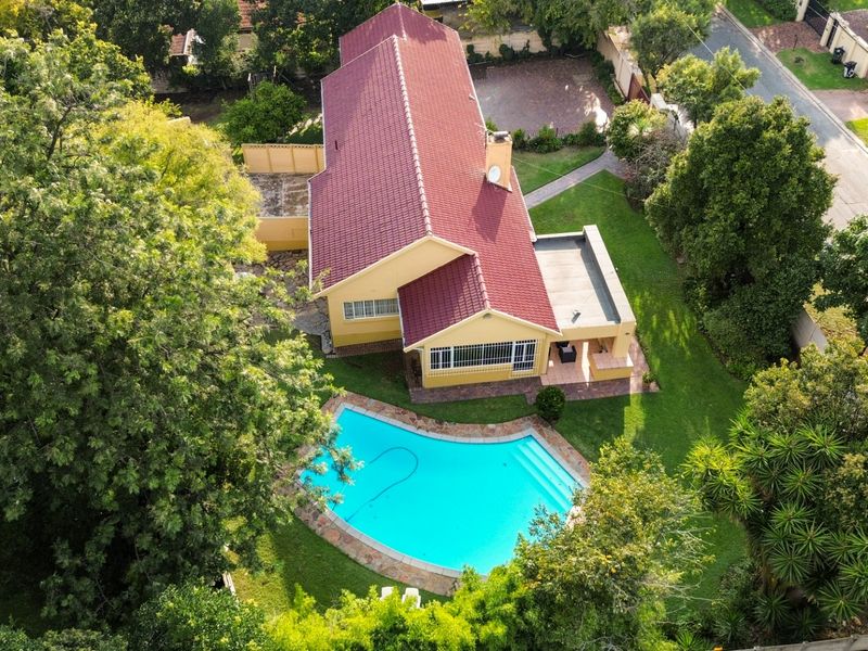 Aerial view of a residential property featuring a large swimming pool, expansive garden, paved patio area, and a well-maintained house with a tiled roof.