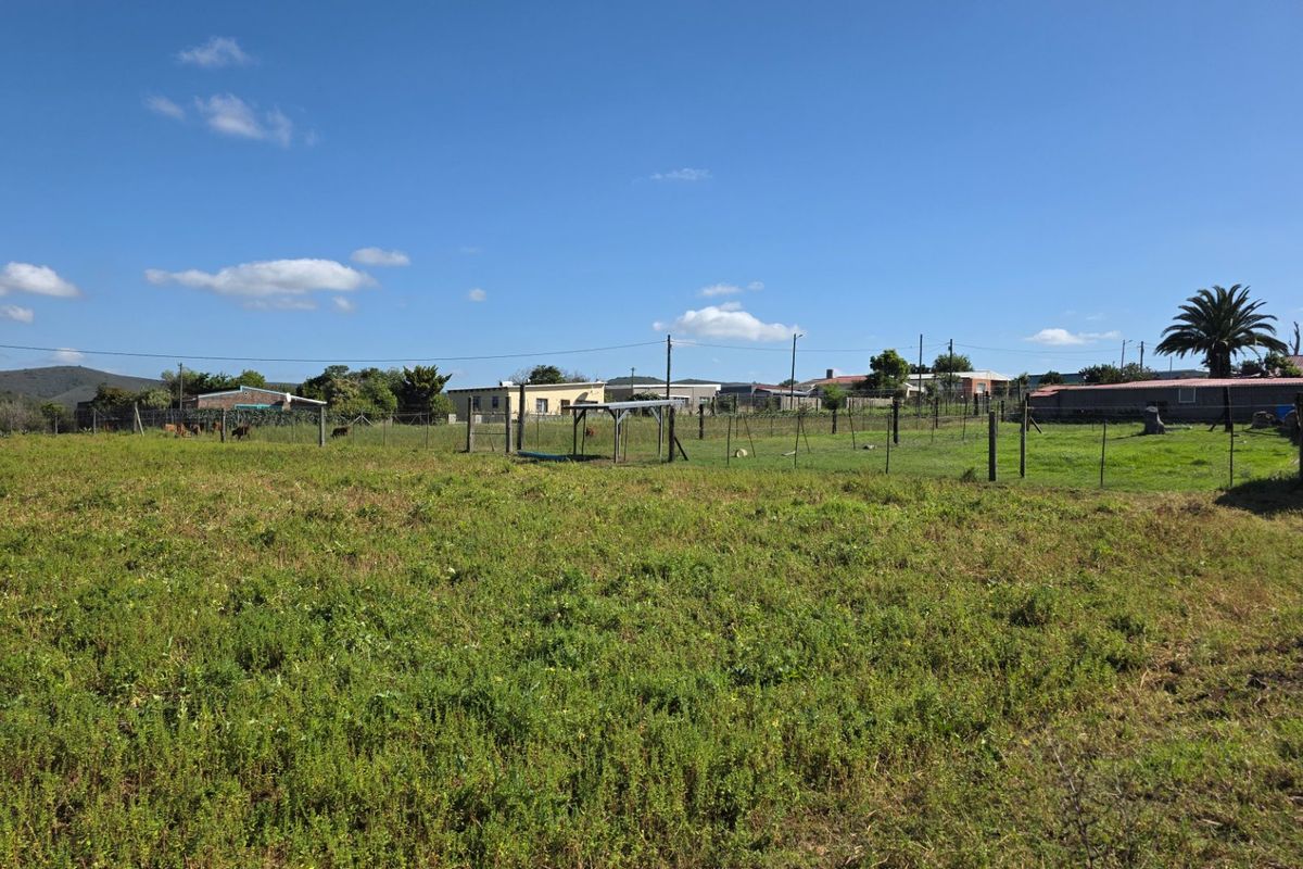 A view of a vacant residential plot featuring open grassy land, perimeter fencing, and surrounding neighborhood structures.