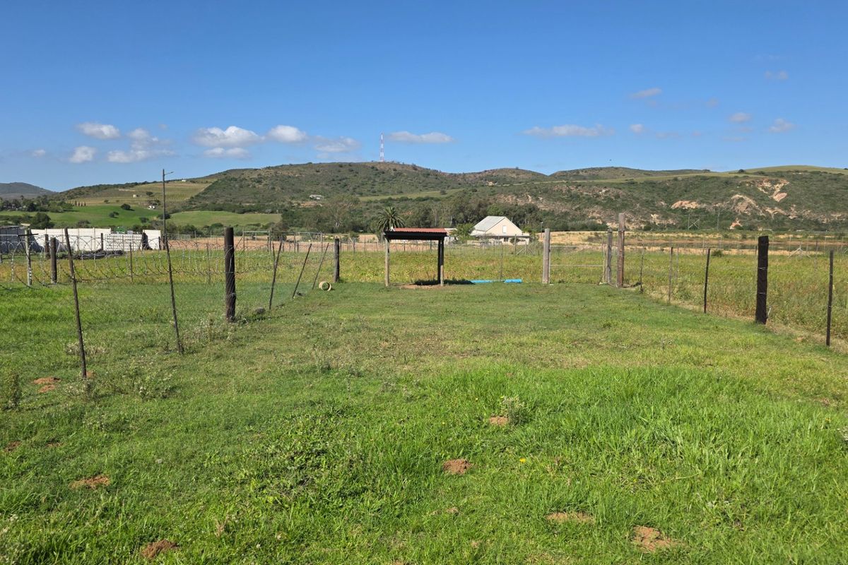 A vacant residential plot featuring open grassy land, perimeter wire fencing, and a small basic shelter structure against a scenic mountain backdrop.