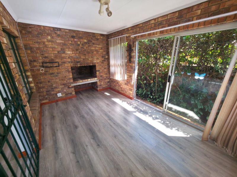 An enclosed patio area featuring exposed brick walls, a built-in brick braai, modern laminate flooring, and large sliding glass doors leading to the garden.