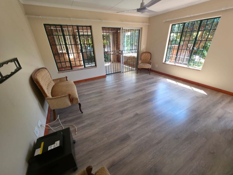 A bright living room featuring modern laminate flooring, a ceiling fan, and large windows with security bars, providing access to the outdoor area.