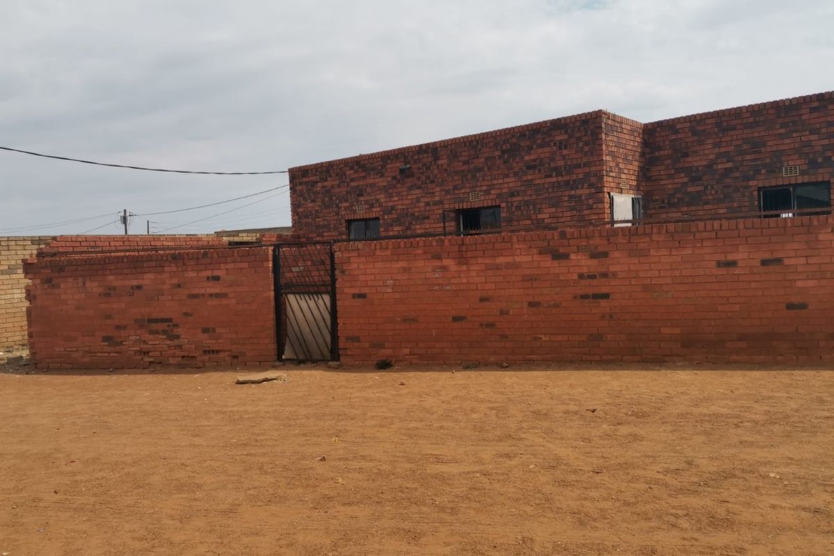 Rear view of a face brick house featuring a perimeter wall with a security gate and unpaved yard area.