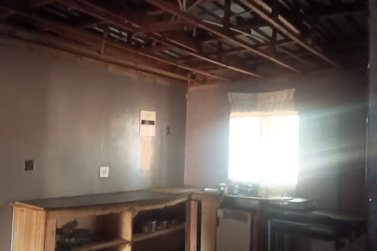 A rustic kitchen space featuring exposed timber roof trusses, built-in wooden cabinetry, and tiled flooring, currently in need of renovation and ceiling installation.