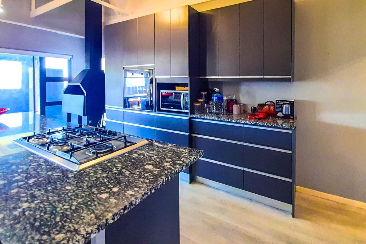 Modern kitchen featuring dark cabinetry, granite countertops, a central island with a gas hob, built-in appliances, and an integrated fireplace.