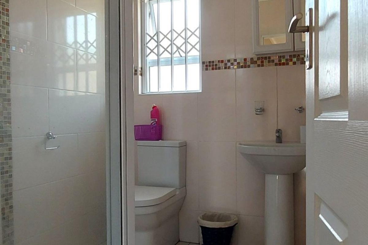 A functional bathroom featuring a glass shower enclosure, pedestal basin, and toilet, finished with neutral wall and floor tiles and security bars on the window.