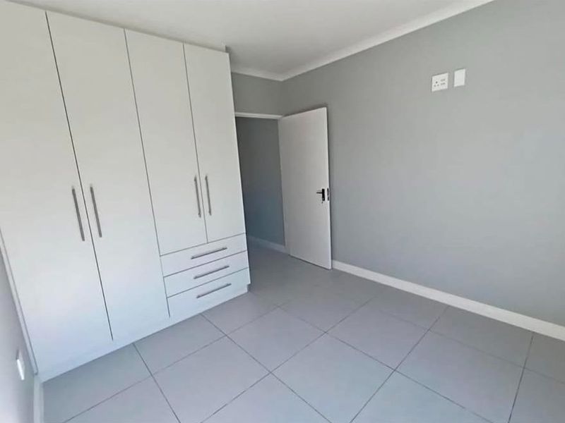 A clean, modern bedroom featuring neutral grey floor tiles, light grey walls, and a large, white built-in wardrobe with ample storage space.
