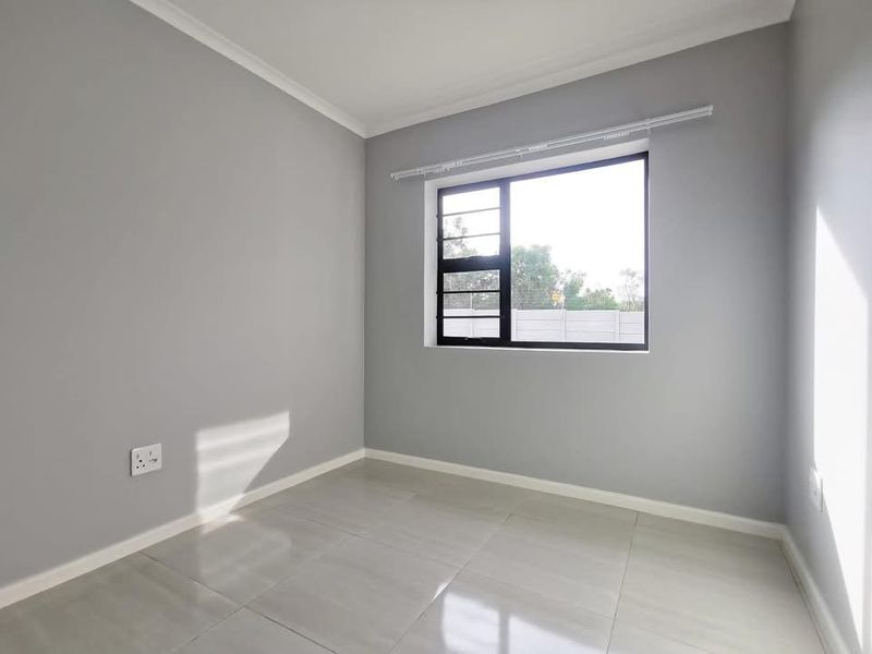 A bright, empty bedroom featuring neutral grey walls, large tiled flooring, and a window with a curtain rail.