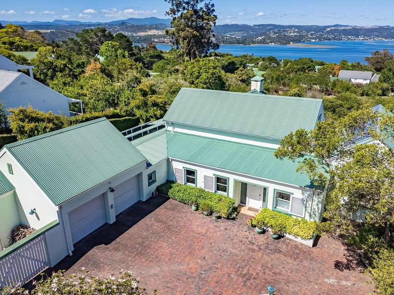 Aerial view of a charming residence featuring a green corrugated metal roof, double garage, and brick-paved driveway, set against a scenic coastal landscape.