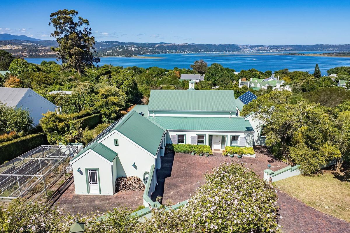Aerial view of the property featuring a green metal roof, solar panels, and a spacious brick-paved driveway, set against a scenic coastal landscape.