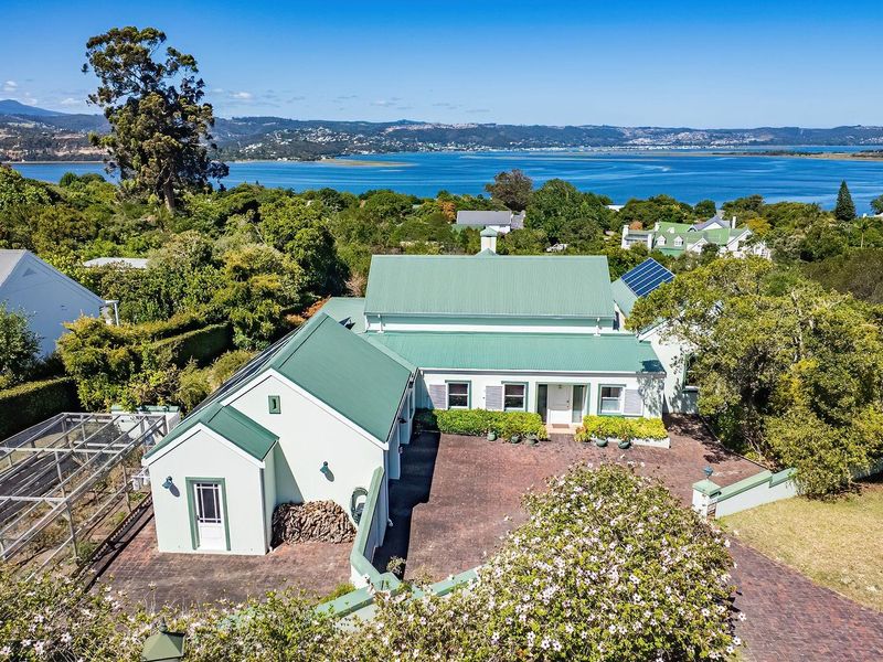Aerial view of the property featuring a green metal roof, solar panels, and a spacious brick-paved driveway, set against a scenic coastal landscape.