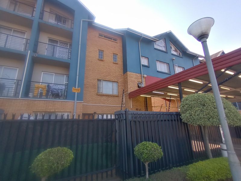Exterior view of a multi-story apartment complex featuring brick and painted facade, metal security fencing, and covered parking area.