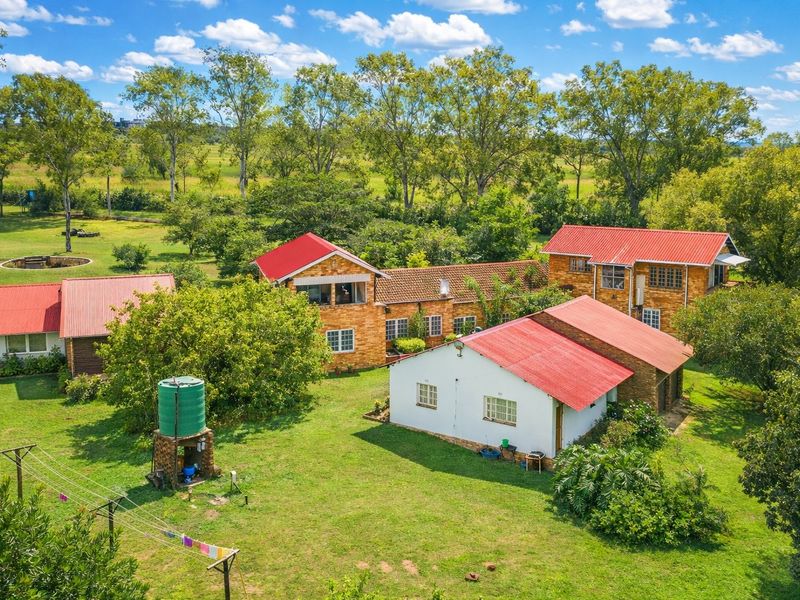 Aerial view of a farm property featuring multiple brick buildings with red corrugated metal roofs, surrounded by expansive green lawns and mature trees.