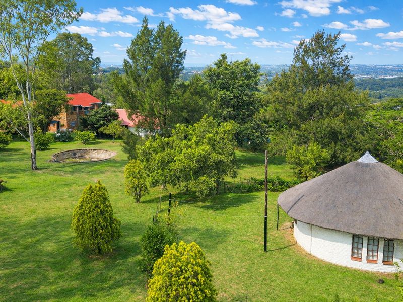 Aerial view of a farm property featuring a thatched-roof structure, a large garden, and an empty swimming pool set against a scenic landscape.