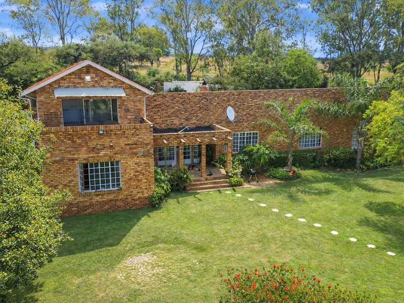 Rear view of a double-story face brick farmhouse featuring a covered patio, established garden, and mature trees in a rural setting.