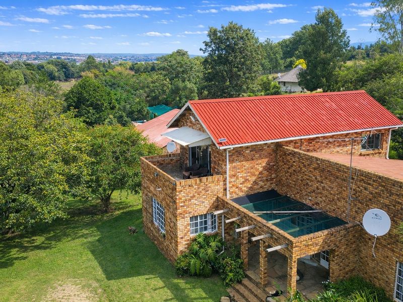 Aerial view of a face brick farmhouse featuring a red corrugated metal roof, a balcony, and a covered patio area with a glass roof section.