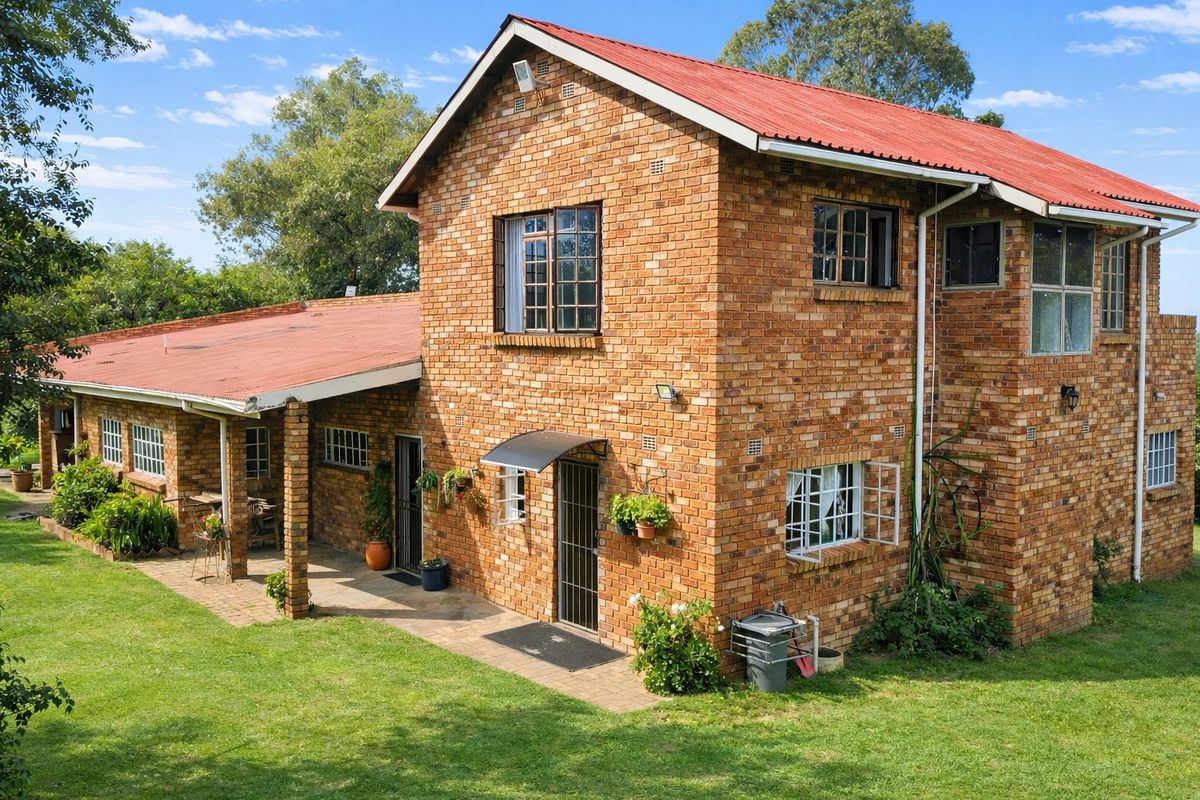 A two-story face brick farmhouse featuring a red corrugated metal roof, covered patio area, and established garden surroundings.
