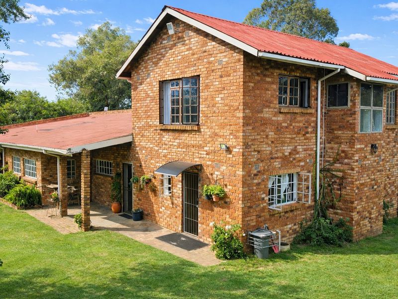 A two-story face brick farmhouse featuring a red corrugated metal roof, covered patio area, and established garden surroundings.