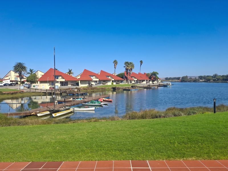 A scenic view of the waterfront featuring a private jetty, manicured lawn, and a row of residential units with red roofs.