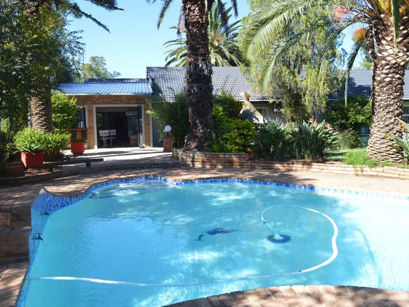 A well-maintained outdoor pool area featuring brick paving, mature palm trees, and a covered patio space attached to the main residence.