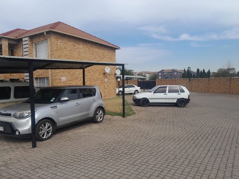 Exterior view of the apartment complex featuring a brick facade, tiled roof, and a paved parking area with covered carports.